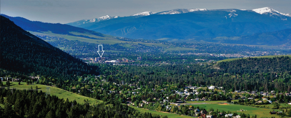 view of Missoula from afar with row of mountains in the background and an arrow pointing out the UM campus