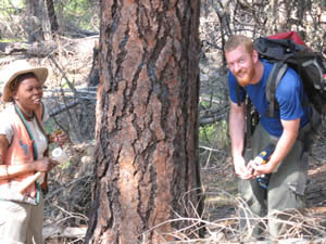 two people posing outside next to the trunk of a conifer tree