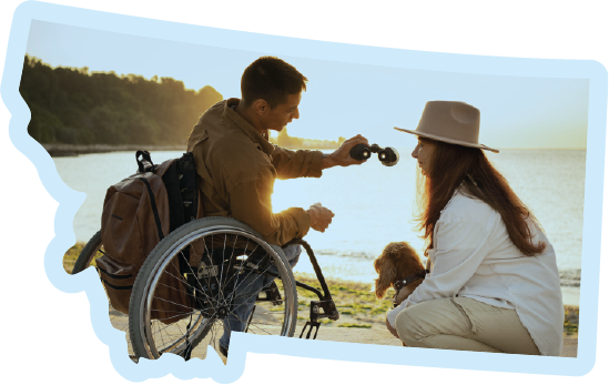 person in wheelchair handing binoculars to another person crouching with dog next to lake
