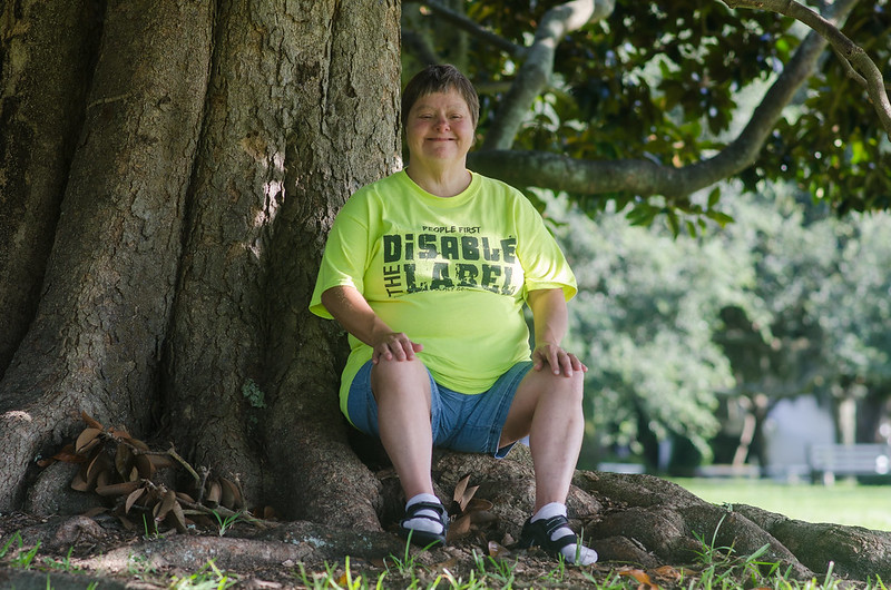 A woman with a disability sits against a tree. Her shirt reads "People first disable the label."