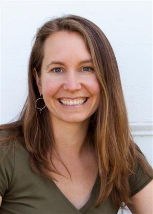 A smiling woman with long brown hair, wearing a green top and earrings, poses against a white background.