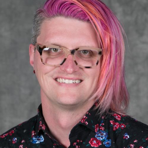 person with pink and orange hair with tortoise shell glasses and roses embroidered on a black shirt.