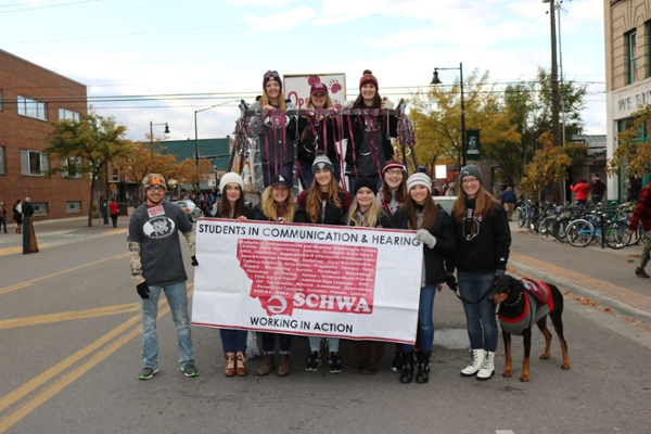 a group of 11 nsslha members standing. Three are in the back row on a truck. The front row is holding a sign taht reads "Students in communication and hearing, working in action."