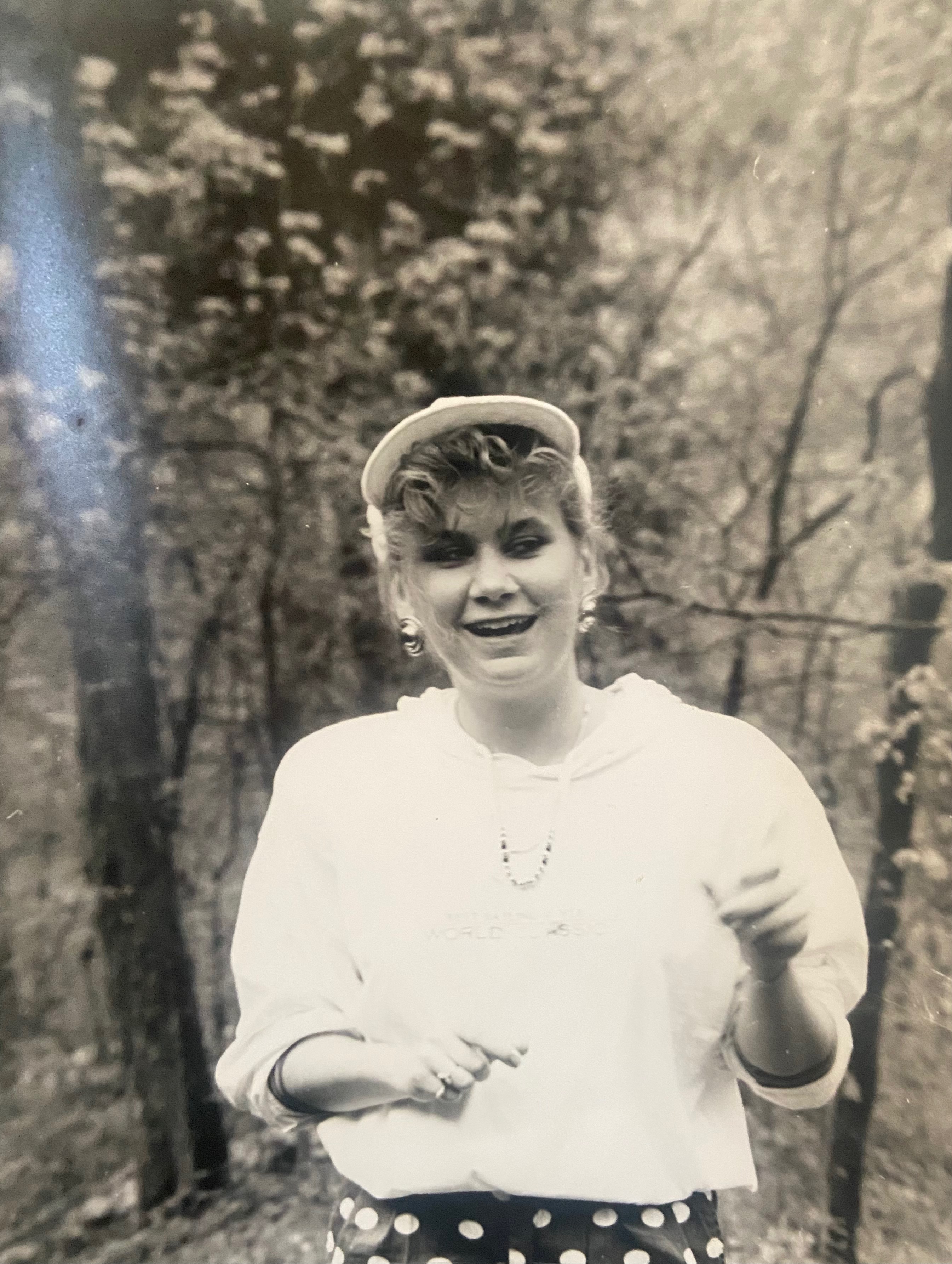 Black and white photo of a smiling woman wearing a cap and a casual outfit, standing outdoors among trees.