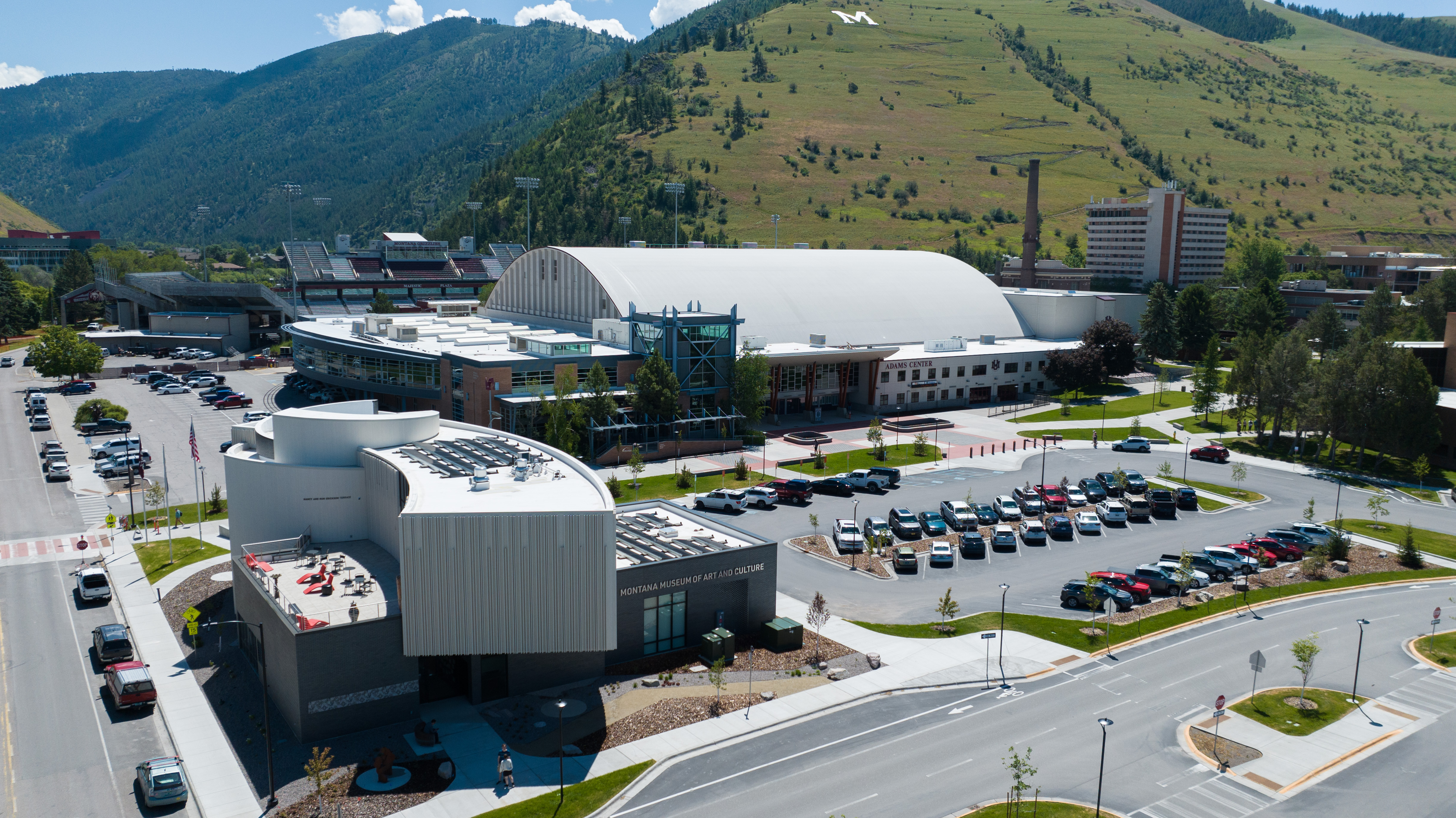 Aerial view of a university campus with the Montana Museum of Art and Culture and the Adams Center with solar panels shown, with mountains in the background.