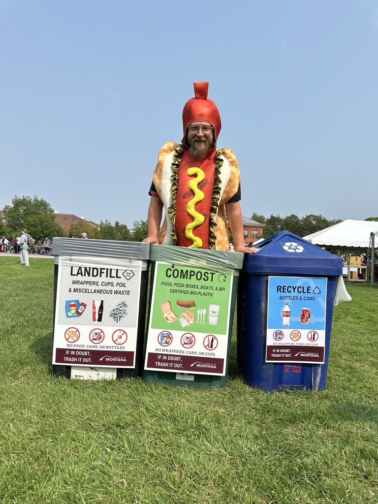 Person in a hot dog costume standing behind landfill, compost, and recycling bins at an outdoor event.