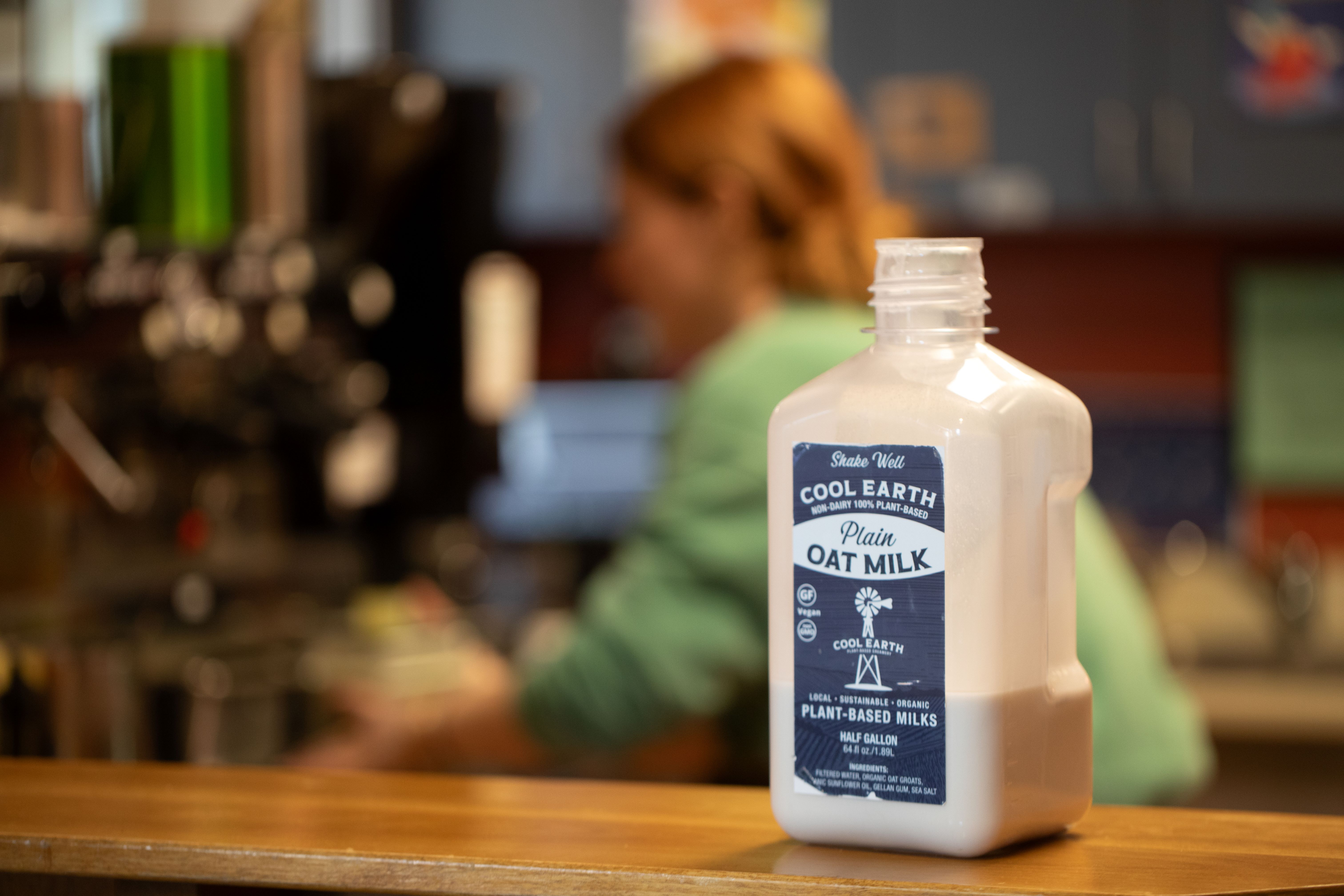 A bottle of Cool Earth plain oat milk sits on a counter with a blurred barista in the background.