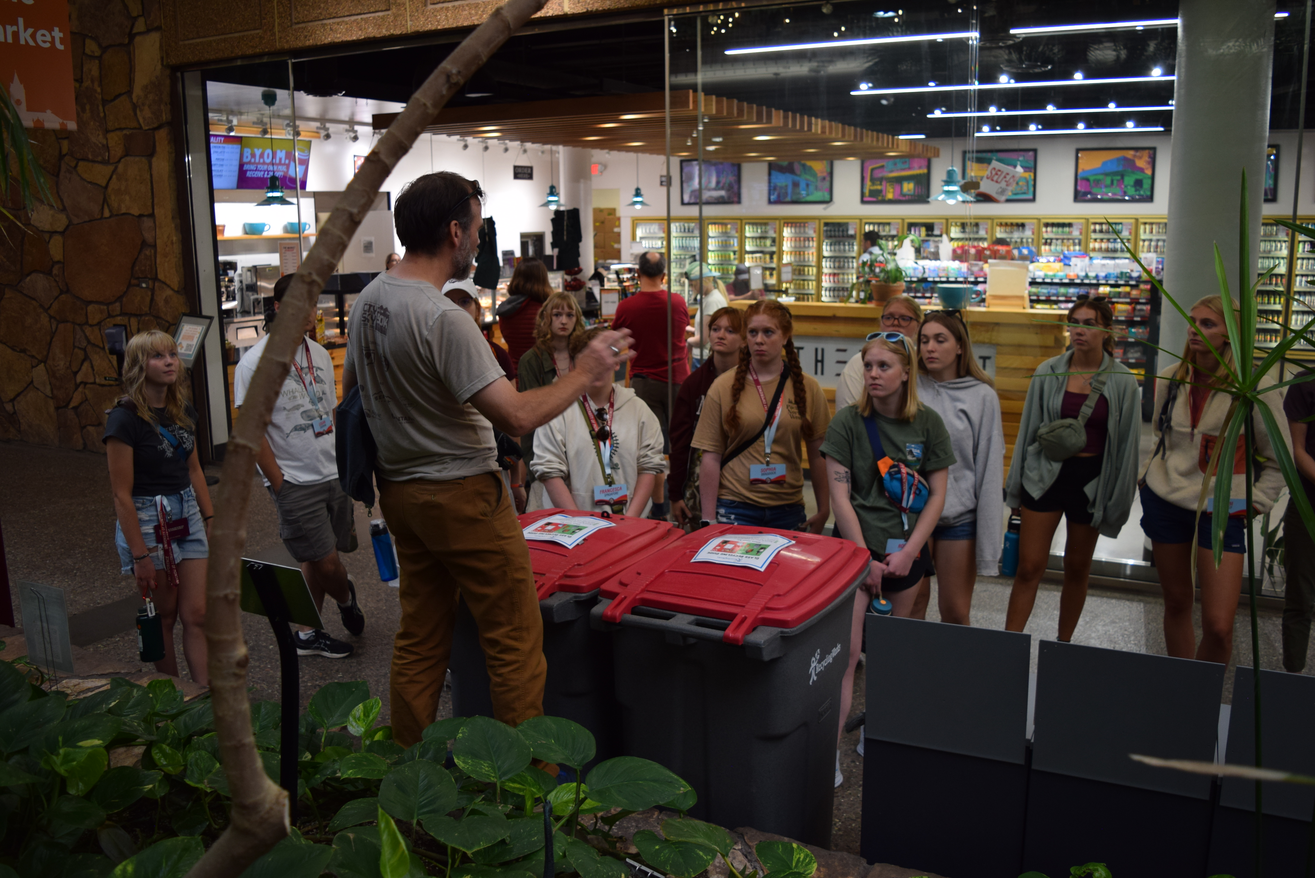 A group of students attentively listening to a person speaking near glass recycling bins outside UC market