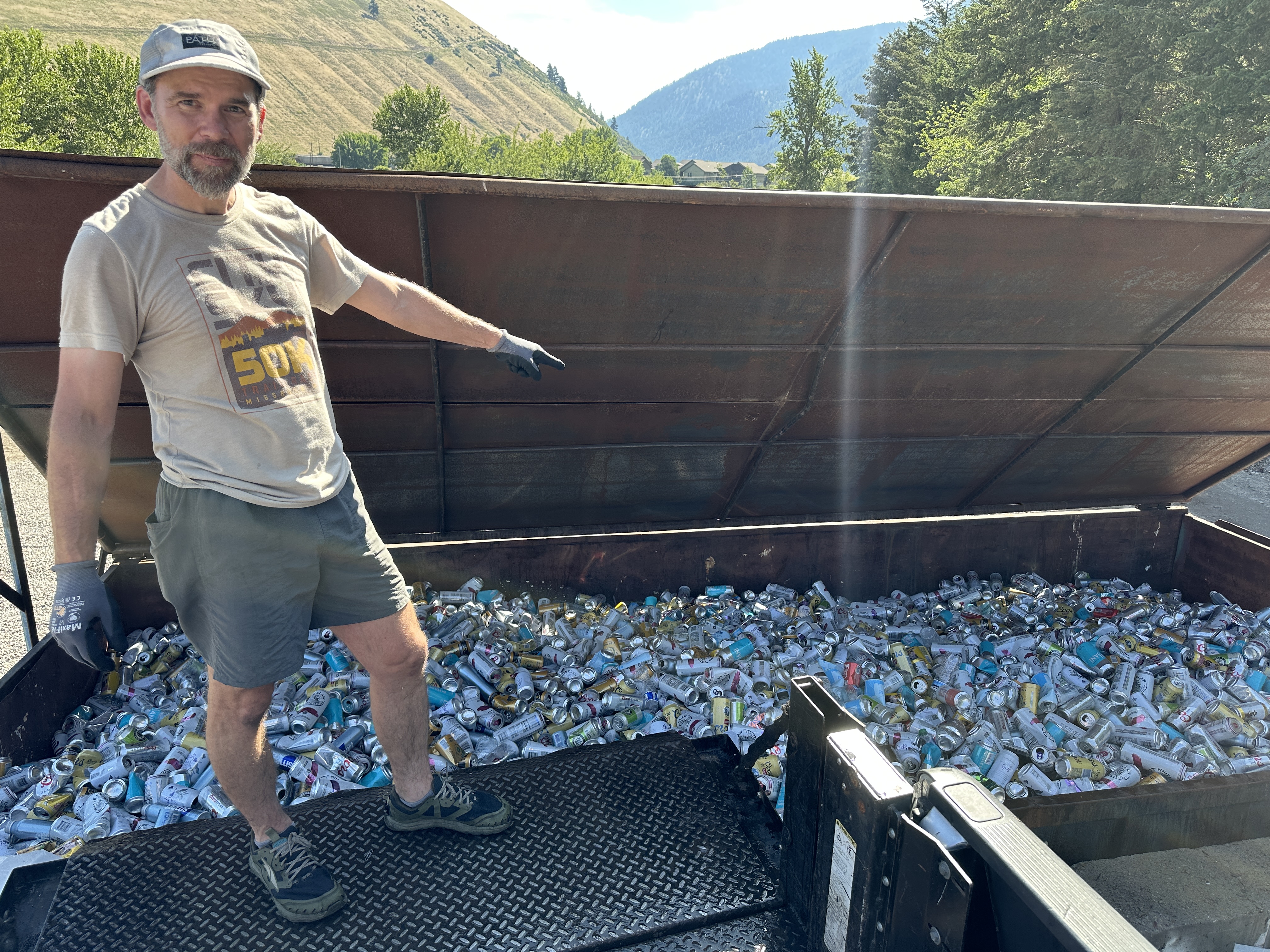 Man standing by a large metal bin filled with crushed aluminum cans, pointing toward the pile with mountains in the background.