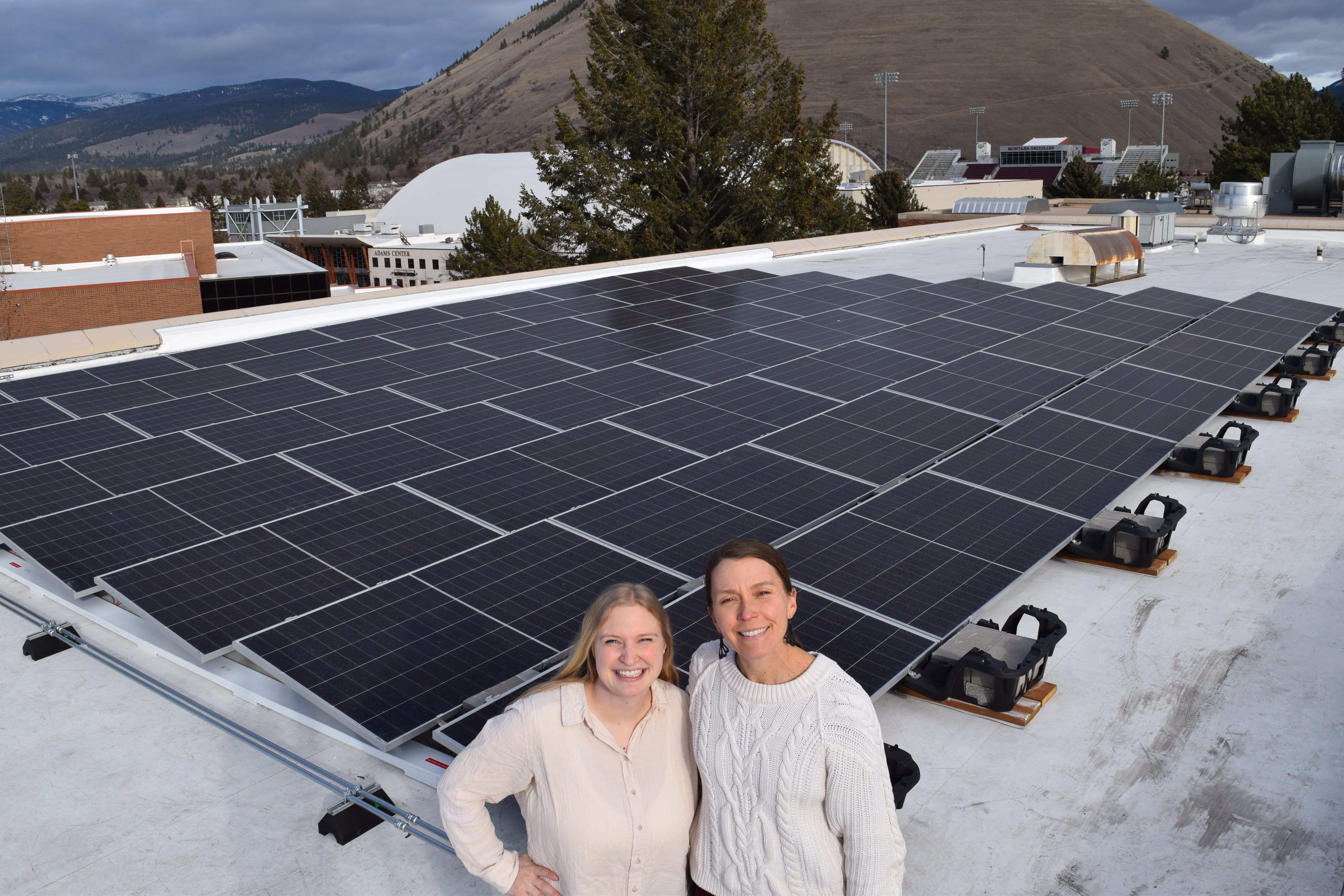 Two people standing on a rooftop next to a large array of solar panels, with mountains in the background.