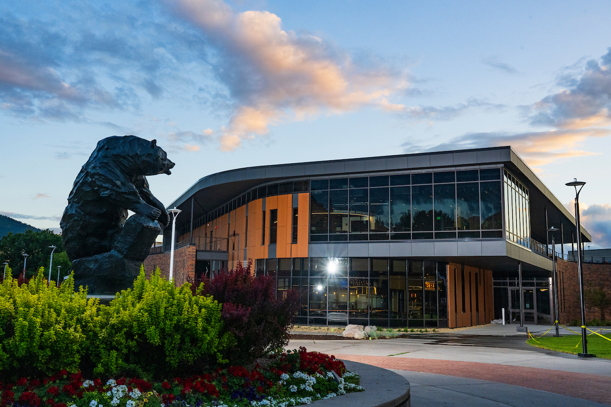 A bear statue stands in front of the lodge dining hall with large glass windows and colorful landscaping.