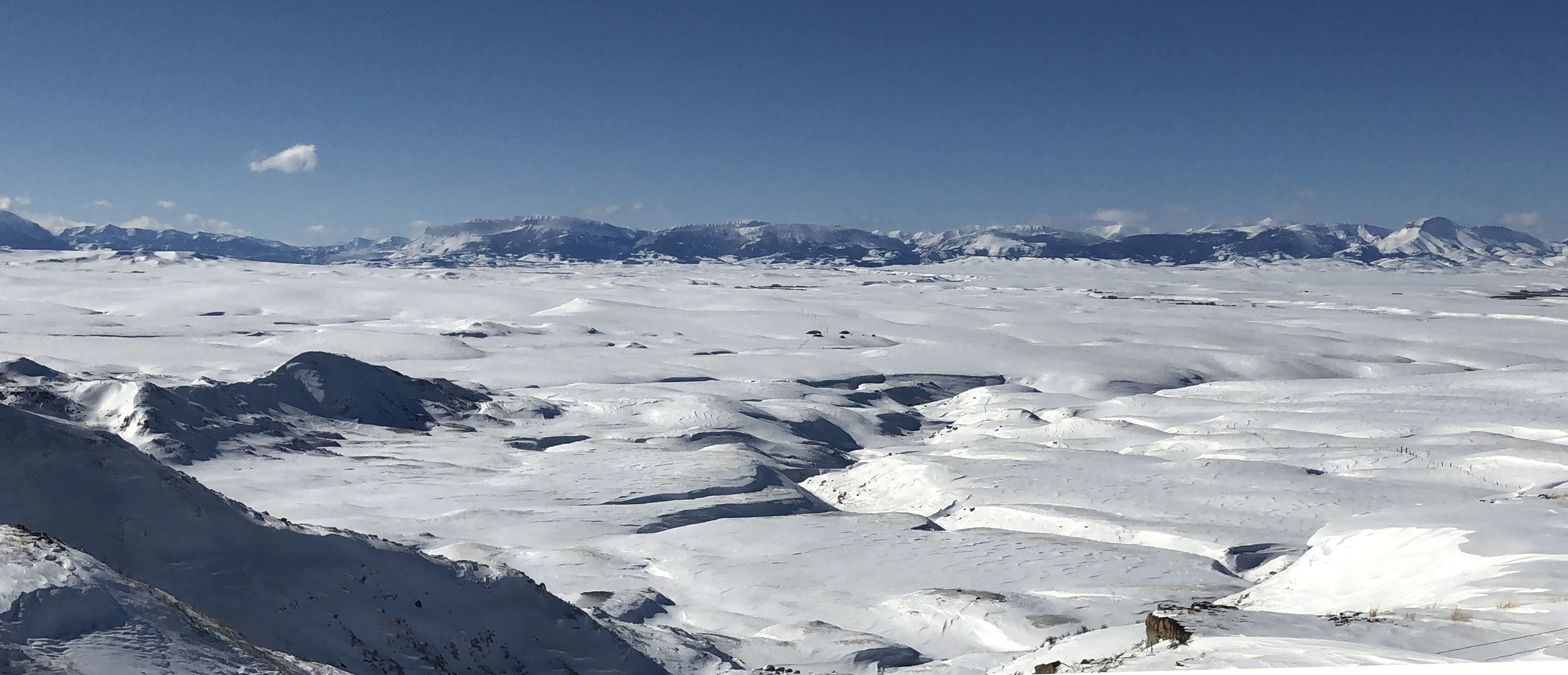Snow covers the rolling landscape in the foreground, while snow-capped mountains protrude in the background on a clear, bluebird day