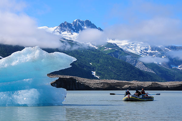 raft being rowing on water next to large iceberg with snowy mountains in the background