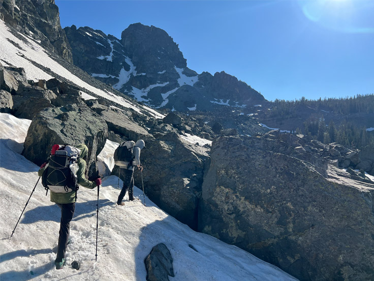 Backpackers hiking through snowy, rocky alpine terrain.