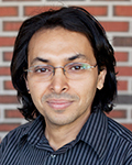 Head-and-shoulders portrait of Khaled Huthaily man wearing glasses and a striped shirt in front of a brick wall.