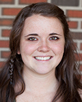 Smiling Hannah Peters with long brown hair standing in front of a brick wall.