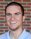 Smiling Brendan Work with short dark hair in a light blue shirt standing in front of a brick wall.