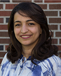 Smiling Amal Al-Misbahi with dark hair wearing a blue-and-white plaid shirt in front of a brick wall.