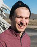 Smiling Jake Green wearing a black beanie and maroon jacket outdoors near a road.
