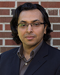 Khaled Huthaily with glasses and dark hair wearing a blazer, standing in front of a brick wall.