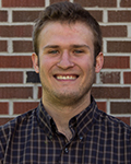 Smiling Royce Mccarty in a plaid shirt standing in front of a brick wall.