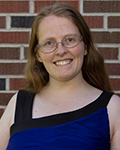 Smiling Susannah Cragwick with glasses in a blue and black top standing in front of a brick wall.