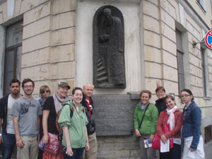 Group of people posing on a street corner beside a stone wall plaque with a relief statue.